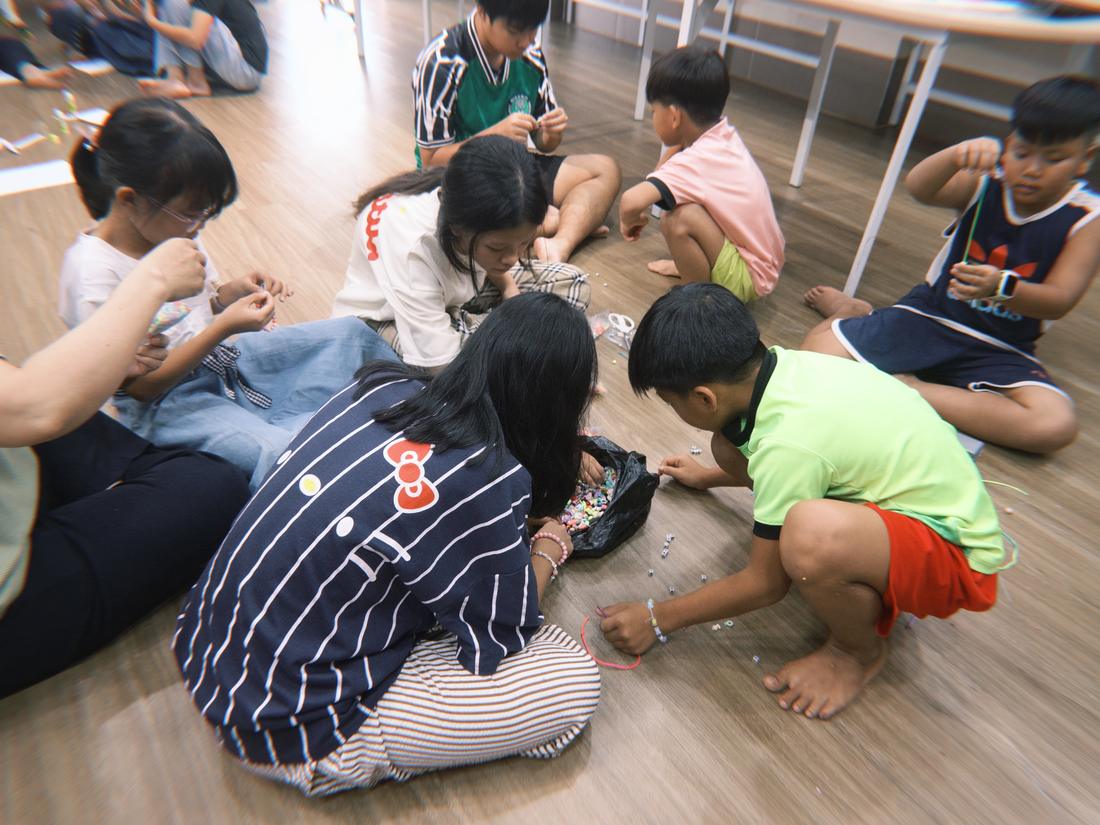 A wide shot of the classroom floor with children spread out making beaded bracelets.